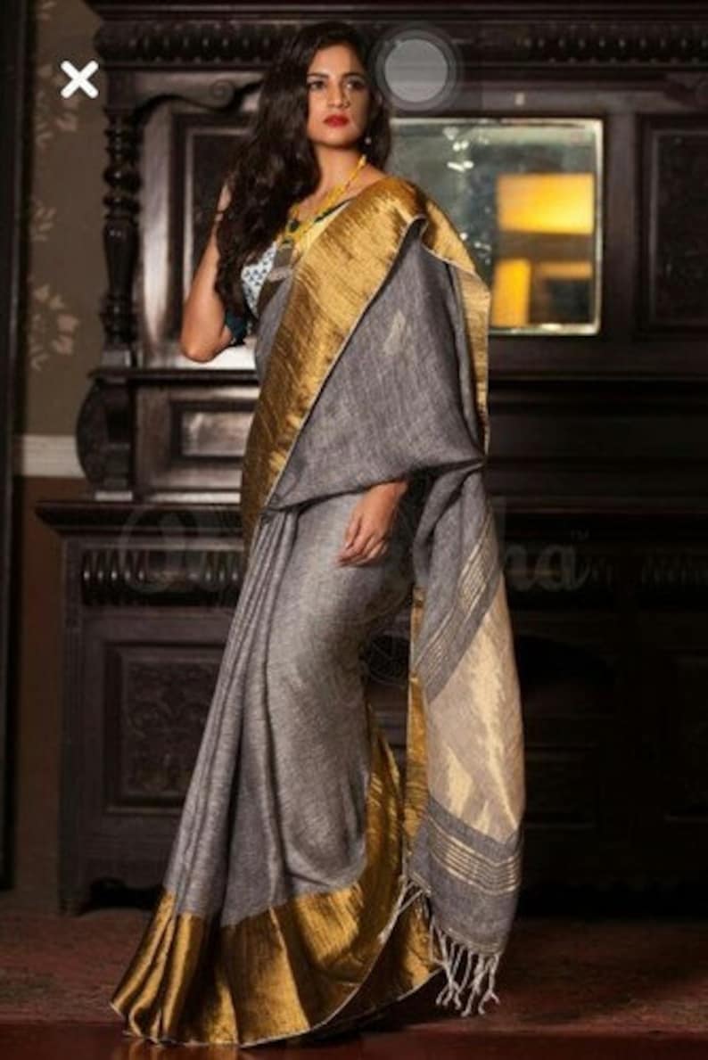 May include: A gray linen saree with a gold border and a white and gold patterned pallu. The saree is draped on a woman standing in front of a dark wooden cabinet.