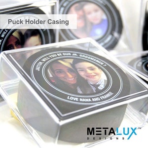 May include: A clear plastic puck holder case with a black hockey puck inside. The puck has a photo of a couple and the text "JAXTON, WILL YOU BE OUR JR. GROOMSMEN? LOVE NANA AND FRANKIE" printed on it.