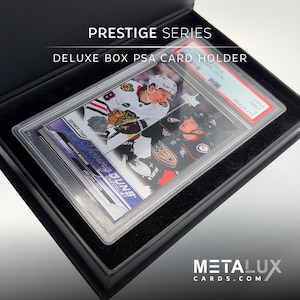 Op de afbeelding: Een zwarte PSA-kaarthouder met een hockeykaart erin. De kaart toont twee hockeyspelers en de tekst "PRESTIGE SERIES DELUXE BOX PSA CARD HOLDER". De kaart zit in een doorzichtige plastic hoes. De website METALUXCARDS.COM is zichtbaar.