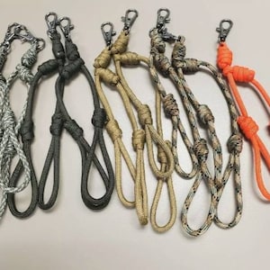 May include: A set of seven colorful paracord lanyards with different patterns and colors, including black, green, tan, camouflage, and orange. Each lanyard has a metal clip at the top and a knot at the bottom.