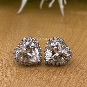May include: A pair of heart-shaped stud earrings. Each earring features a large, clear, heart-cut stone surrounded by a halo of smaller, sparkling stones. The earrings are set in a silver-colored metal.