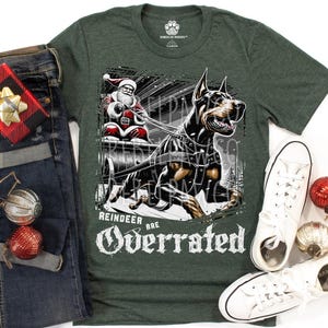 May include: A dark green t-shirt featuring a graphic of a Doberman dog pulling a sleigh with Santa Claus. The text "REINDEER ARE OVERRATED" is printed in white below the graphic. The shirt is displayed alongside blue jeans and white sneakers.