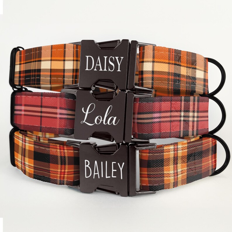 May include: Three dog collars with plaid patterns in shades of orange, red, and brown. Each collar has a black buckle with a name: "Daisy", "Lola", and "Bailey". The collars have black D-rings.
