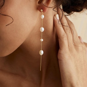 May include: A pair of long, delicate gold earrings with a chain and five white pearls. The pearls are different sizes and are evenly spaced along the chain.
