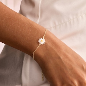 May include: A delicate gold-toned bracelet featuring a daisy charm with white petals and a yellow center. The bracelet is worn on a wrist against a white fabric background. The daisy charm is approximately 0.5 inches in diameter.