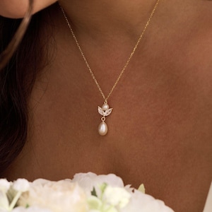 May include: A delicate gold necklace featuring a small pendant with a pearl drop. The pendant has a cluster of small, clear crystals and a small pearl at the top. The necklace is worn on a person's neck, with a bouquet of white flowers in the foreground.