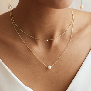 Peut inclure: Un collier délicat en chaîne dorée avec un pendentif en perle unique. Le collier est superposé à une chaîne plus courte avec une petite perle dorée.