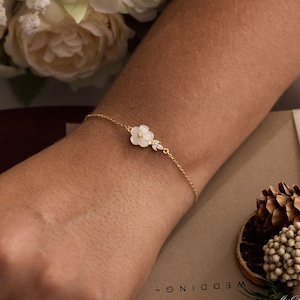 May include: A delicate gold bracelet featuring a white flower and small, sparkling accents. The bracelet is worn on a wrist, with a blurred background of flowers and wedding-themed stationery.