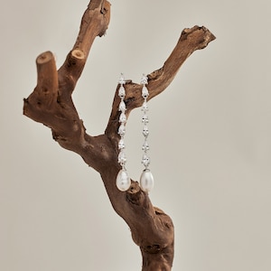 May include: A pair of long, silver earrings with pear-shaped crystals and a pearl drop at the bottom. The earrings are hanging from a piece of driftwood.