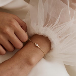 May include: A delicate gold chain bracelet with a single pearl charm. The bracelet is shown on a person's wrist.