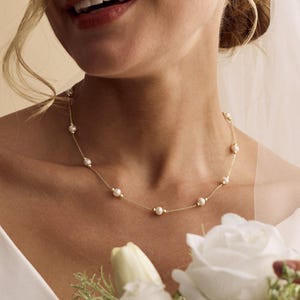 Könnte beinhalten: Eine zarte goldene Halskette mit runden, weißen Perlen. Die Halskette wird von einer Person in einem weißen Kleid getragen, die einen Strauß weißer Blumen hält. Ein schlichtes, elegantes Schmuckstück.
