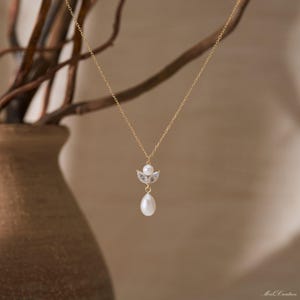 May include: A delicate gold necklace featuring a pendant with a small pearl, two clear crystals, and a larger teardrop-shaped pearl. The necklace hangs against a soft, neutral background, highlighting the jewelry's elegant design.