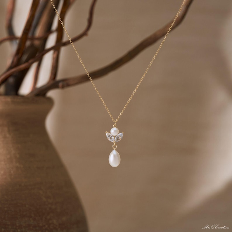 May include: A delicate gold necklace featuring a pendant with a small pearl, two clear crystals, and a larger teardrop-shaped pearl. The necklace hangs against a soft, neutral background, highlighting the jewelry's elegant design.
