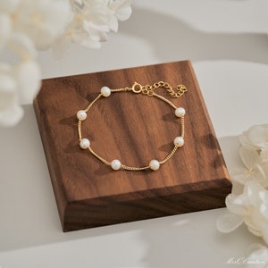 May include: A delicate gold chain bracelet with small white pearls. The bracelet is displayed on a wooden surface.