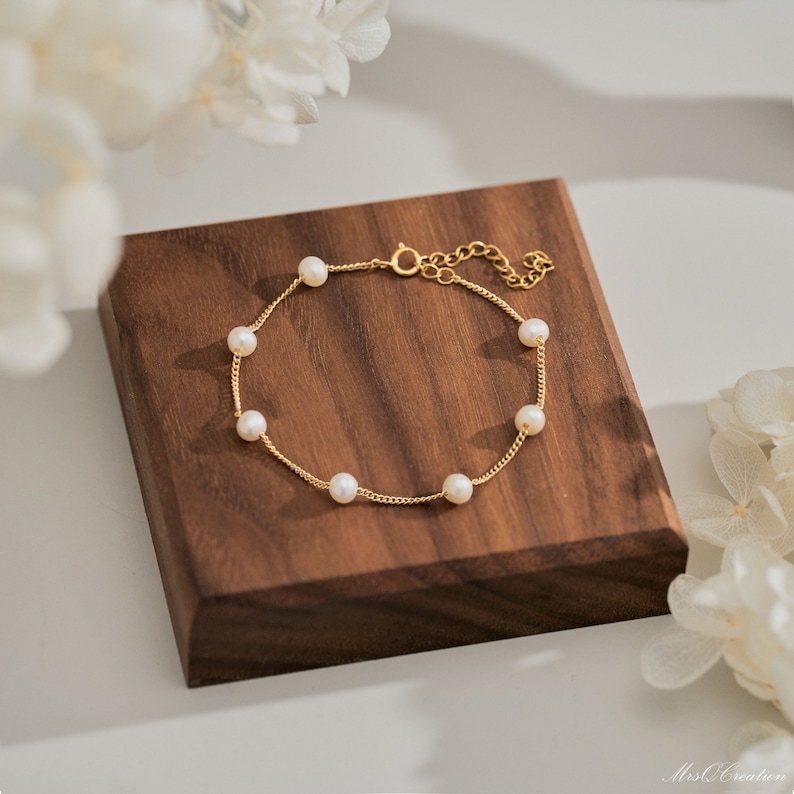 May include: A delicate gold chain bracelet with small white pearls. The bracelet is displayed on a wooden surface.