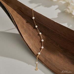 May include: A delicate gold chain bracelet with small white pearls. The bracelet is on a brown wooden surface.