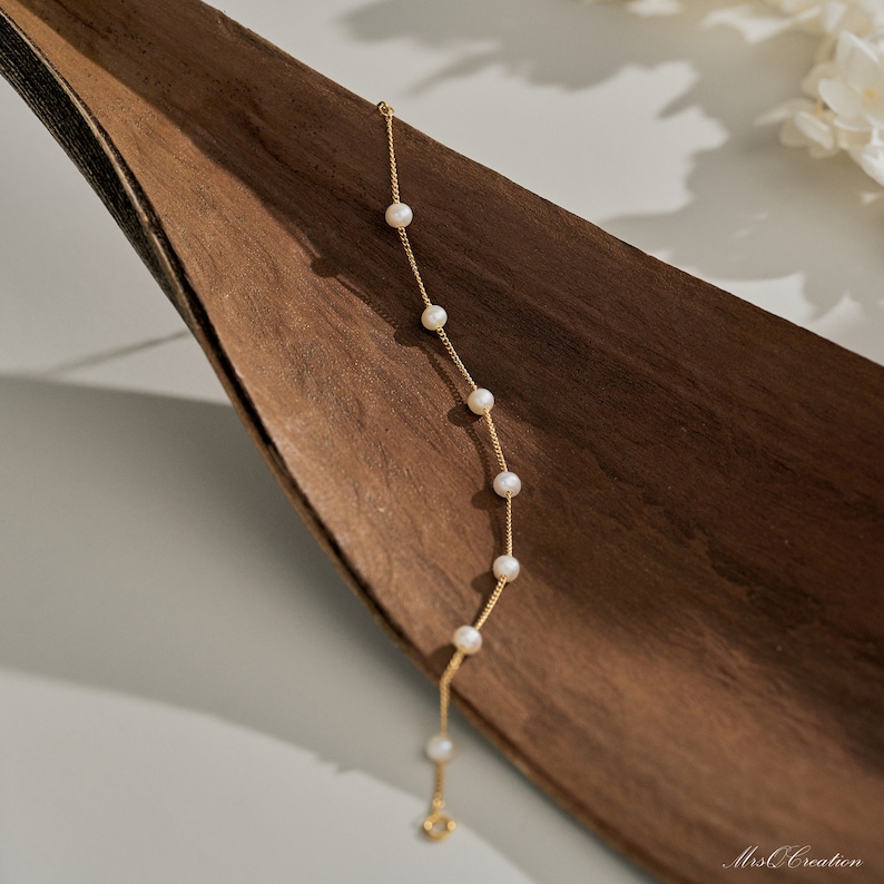 May include: A delicate gold chain bracelet with small white pearls. The bracelet is on a brown wooden surface.