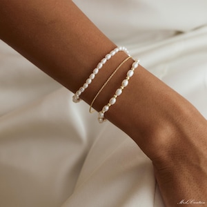 May include: Three bracelets on a wrist. The top bracelet is a pearl strand, the middle bracelet is a delicate gold chain, and the bottom bracelet is another pearl strand.