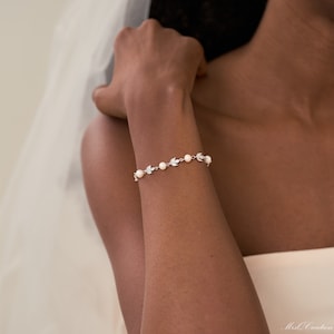 Könnte beinhalten: Ein zartes silbernes Armband mit kleinen weißen Perlen und blattförmigen Anhängern. Das Armband befindet sich an einem Handgelenk.