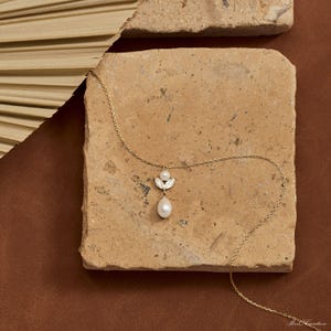 May include: A delicate gold necklace featuring a pendant with a small pearl and a larger, teardrop-shaped pearl. The pendant is accented with small, sparkling embellishments. The necklace is displayed on a textured, light brown surface.