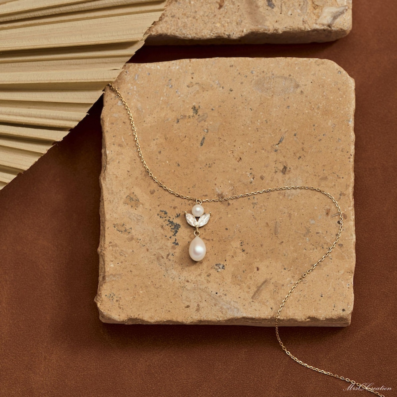 May include: A delicate gold necklace featuring a pendant with a small pearl and a larger, teardrop-shaped pearl. The pendant is accented with small, sparkling embellishments. The necklace is displayed on a textured, light brown surface.
