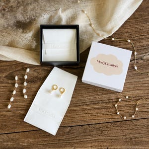 May include: A white box with the text "MrsQCreation" in a cloud shape. The box is on a wooden surface with a white fabric and pearl jewelry.