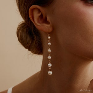 Puede incluir: Pendientes largos dorados con una serie de perlas blancas suspendidas de un delicado gancho. Las perlas varían en tamaño, creando un efecto de cascada. Los pendientes se muestran en una oreja, destacando su longitud y diseño.