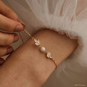 May include: A delicate gold chain bracelet with a pearl and two crystal leaf charms. The bracelet is on a person's wrist.