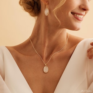 May include: A gold necklace with a white oval pendant and matching earrings. The necklace and earrings are on a model wearing a white shirt.