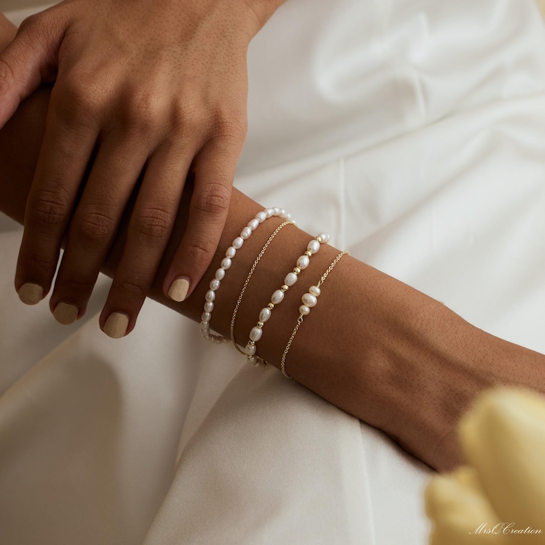 May include: A set of four pearl bracelets on a person's wrist. The bracelets are made of small, white pearls and gold chain. The bracelets are stacked on top of each other.