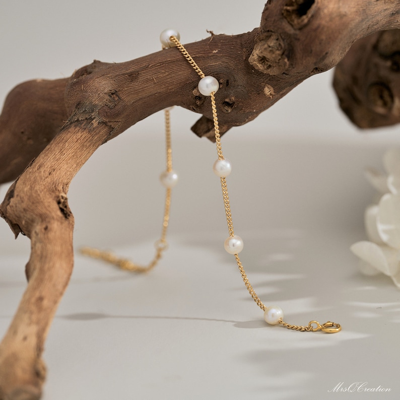 May include: A delicate gold chain necklace with five white pearls. The necklace is draped over a piece of driftwood.