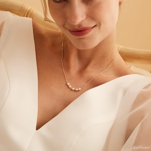 May include: A delicate gold necklace with a row of small, white pearls. The necklace is worn by a person with a white top.