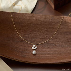 May include: A delicate gold necklace with a pendant featuring a white pearl and two smaller pearls. The pendant is set with small, clear crystals. The necklace is displayed on a wooden surface.