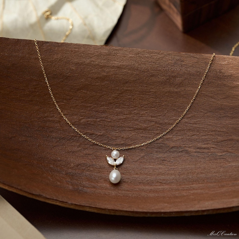 May include: A delicate gold necklace with a pendant featuring a white pearl and two smaller pearls. The pendant is set with small, clear crystals. The necklace is displayed on a wooden surface.