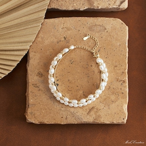 May include: A delicate gold chain bracelet with two rows of white pearl beads. The bracelet is on a light brown surface.