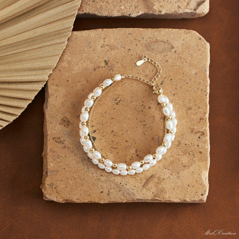 May include: A delicate gold chain bracelet with two rows of white pearl beads. The bracelet is on a light brown surface.
