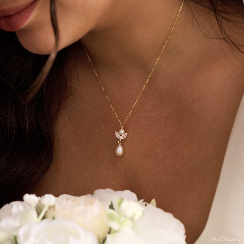 May include: A delicate gold necklace featuring a small pendant with a pearl drop. The pendant has a cluster of small, clear crystals and a small pearl at the top. The necklace is worn on a person's neck, with a bouquet of white flowers in the foreground.
