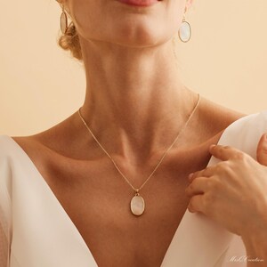 May include: A gold chain necklace with a white oval pendant. The pendant has a smooth, slightly textured surface. The necklace is worn by a person with a white shirt.