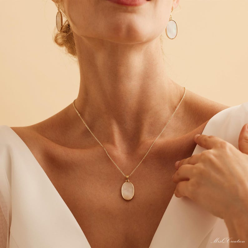 May include: A gold chain necklace with a white oval pendant. The pendant has a smooth, slightly textured surface. The necklace is worn by a person with a white shirt.