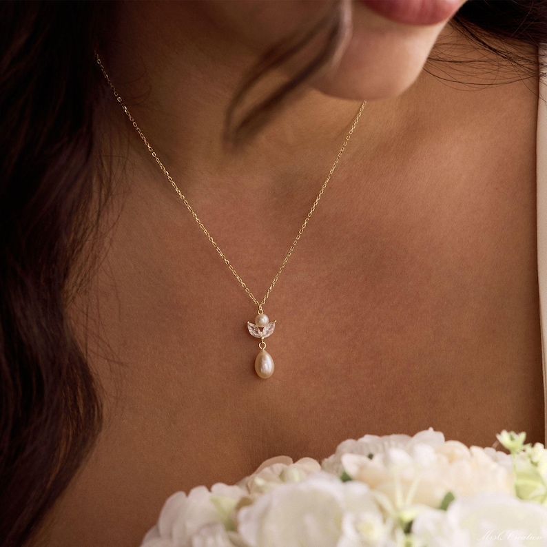 May include: A delicate gold necklace featuring a small, ornate pendant with a teardrop pearl. The pendant has a floral design with small, clear crystals. The necklace is worn against a skin tone background, with a bouquet of white flowers in the foreground.
