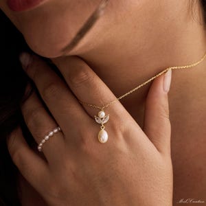 May include: A delicate gold necklace with a pendant featuring a teardrop pearl and small clear crystals. A matching pearl ring is on the finger. The jewelry is set against a soft, blurred background.