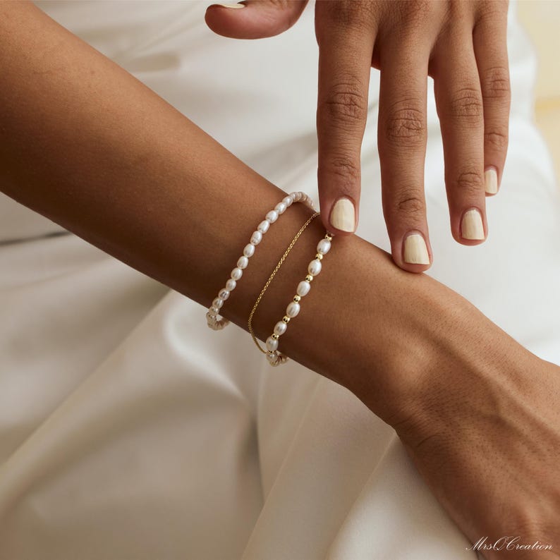 May include: A set of three bracelets on a person's wrist. The bracelets are made of small white pearls and a thin gold chain.