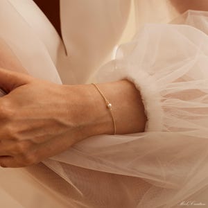 May include: A delicate gold chain bracelet with a single pearl charm. The bracelet is worn on a person's wrist.