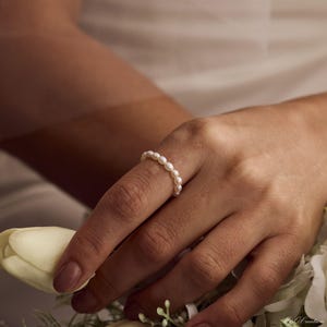 Puede incluir: Un delicado anillo con una fila continua de pequeñas perlas blancas. El anillo se lleva en un dedo, mostrando su elegante diseño. Las perlas están espaciadas uniformemente, creando un aspecto clásico y atemporal. El anillo es una joya.