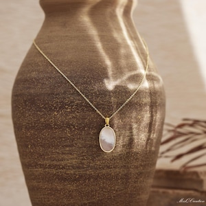 May include: A gold chain necklace with a white oval pendant. The pendant has a pearlescent finish and is set against a brown background.