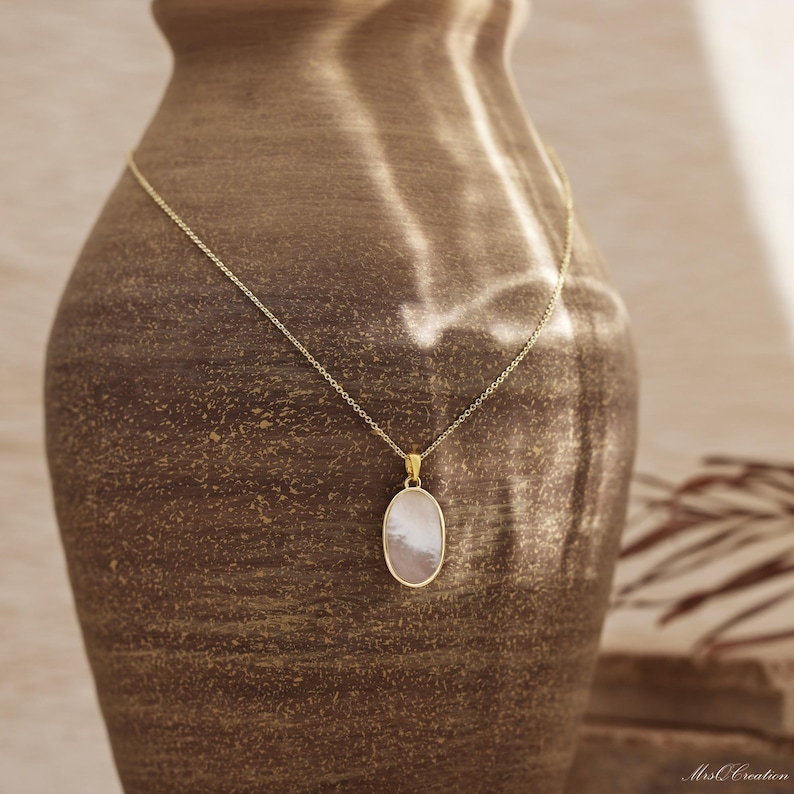 May include: A gold chain necklace with a white oval pendant. The pendant has a pearlescent finish and is set against a brown background.