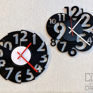 May include: Two black vinyl record clocks with silver numbers and red and white hands. The clock on the left has the numbers arranged in a circle, while the clock on the right has the numbers arranged in a scattered pattern.