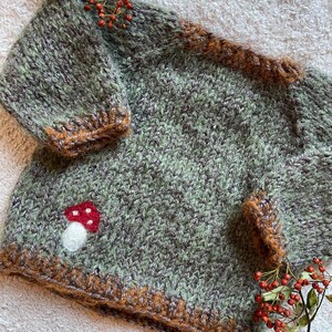 Könnte beinhalten: Ein handgestrickter Pullover aus meliertem salbeigrünem und braunem Garn mit flauschiger Textur. Das Kleidungsstück hat einen Rundhalsausschnitt, lange Ärmel und ein kleines, nadelgefilztes rot-weißes Pilzmotiv auf der unteren linken Vorderseite.