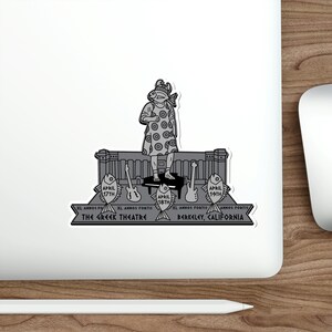 Puede incluir: Una pegatina en blanco y negro que presenta una ilustración estilizada de una figura con cabeza de pez y un vestido estampado, de pie en un escenario. La pegatina incluye el texto "The Greek Theatre, Berkeley, California" y las fechas: 17, 18 y 19 de abril.
