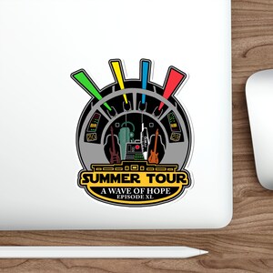 Puede incluir: Una pegatina con un diseño estilizado de cabina de nave espacial, guitarras y el texto "SUMMER TOUR A WAVE OF HOPE EPISODE XL". El diseño incluye acentos verdes, amarillos, azules y rojos.